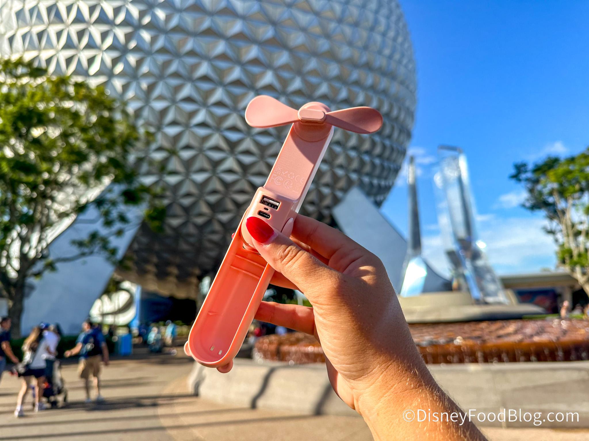 A handheld fan from the JISULIFE brand, held in front of the iconic Spaceship Earth at Epcot in Walt Disney World.